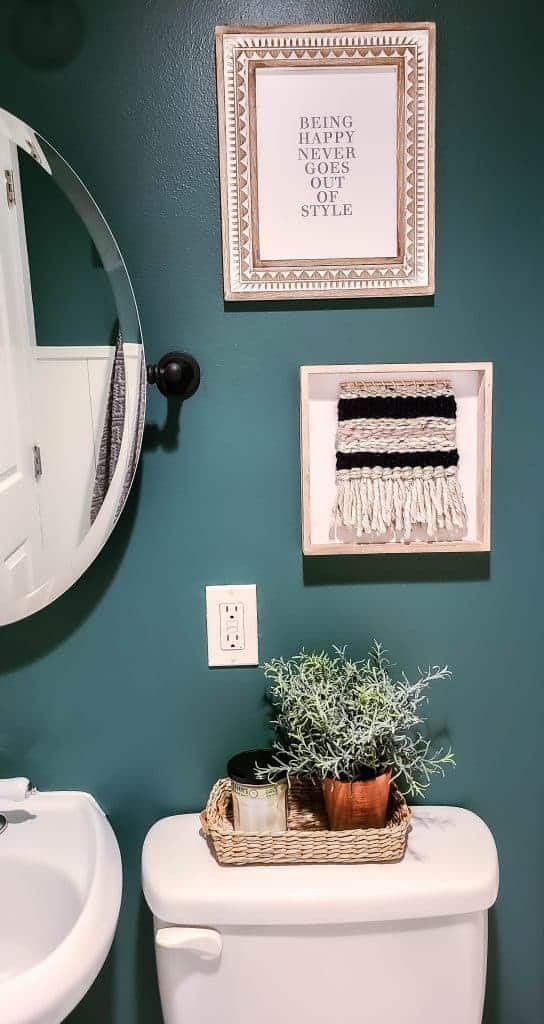 Two pieces of wall art we hung on out bathroom's new deep green walls. One picture is a wooden frame with geometric patterns, and white paper with the text "being happy never goes out of style." The second piece of wall art is woven fabric framed in a simple wooden frame.