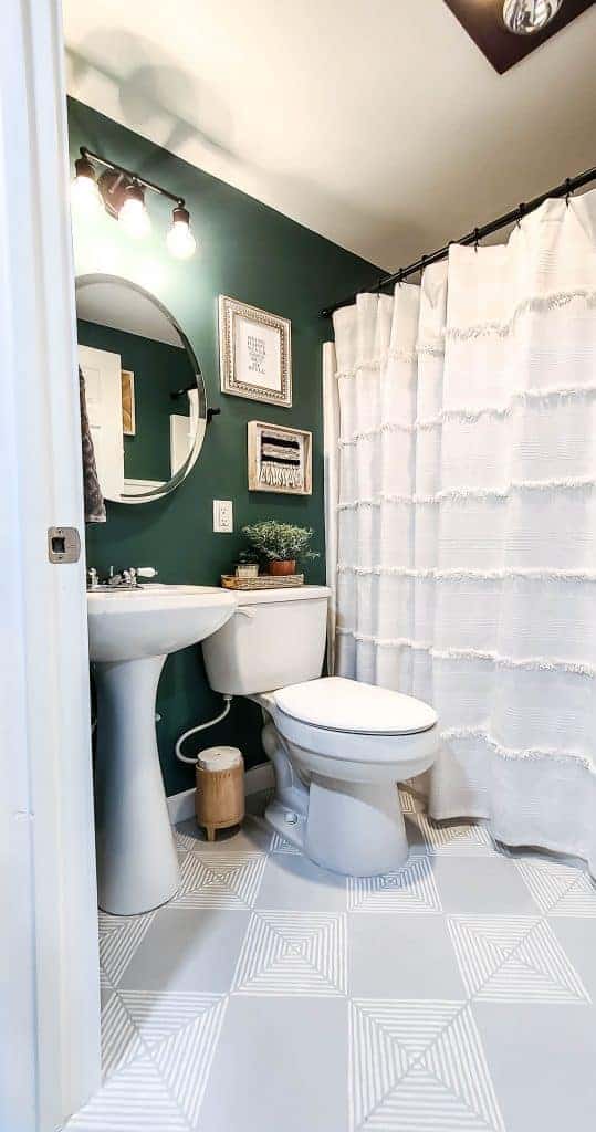 An inside look out our new Buget Bathroom Makeover - deep green painted walls decorated with bohemian style wall art, a white linen shower curtain, and round vanity mirror.