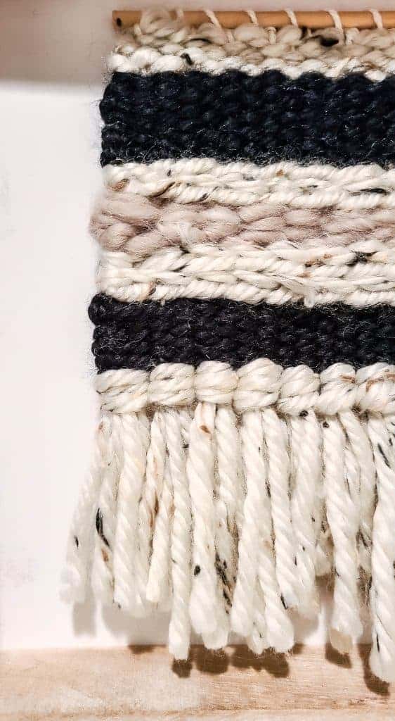 A close up look of a boho style woven wood wall hanging, made up of tan, black, and light brown woven wool against a white background.