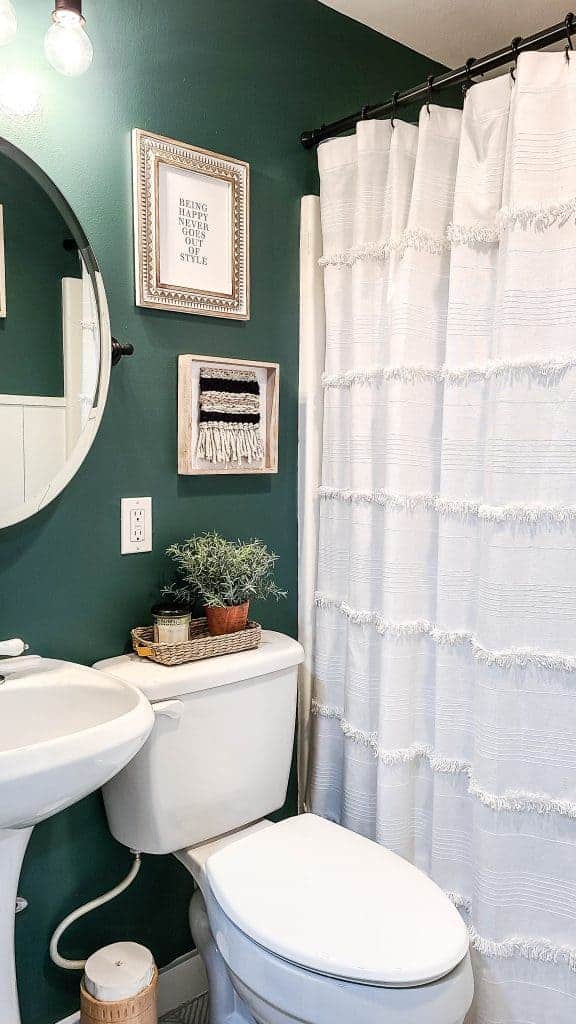 A closer look at the white linen shower curtain, boho style wall decor, green painted walls, and white toilet in our guest bathroom.