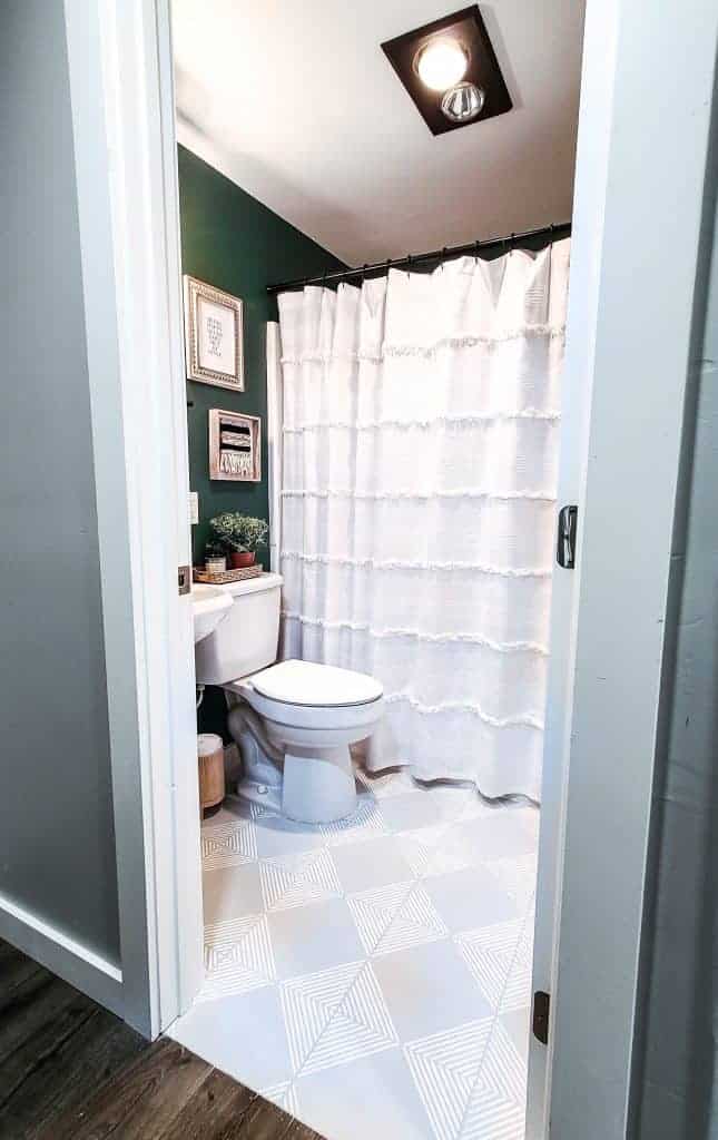 A look inside our updated guest bathroom - an updated grey and white tile floor with a stenciled geometric pattern, white linen shower curtain, deep green painted walls and boho chic wall decor.