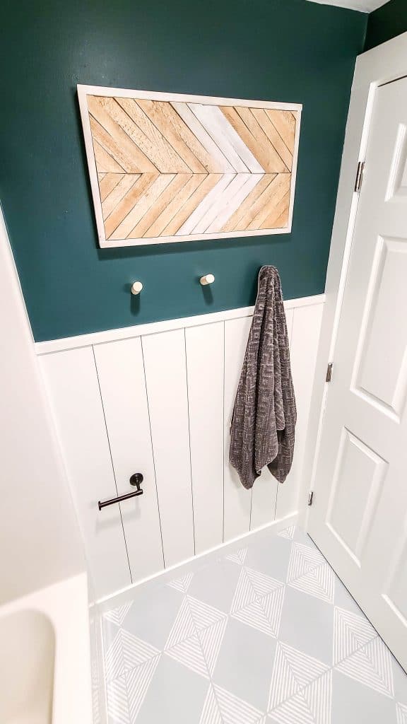 We created a fun two toned bathroom wall with DIY white shiplap on the bottom half, deep green paint on the top half. A DIY wood wall art hangs on the wall, and we see the grey and white tile floor.