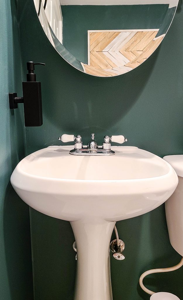 A close up look at the free standing white sink, round glass vanity mirror hanging on the deep green wall, and a wall mounted soap dispenser.