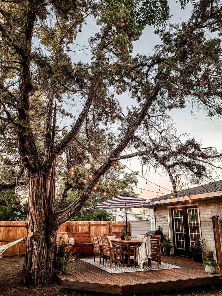 The view of our cozy patio space from the backyard - a wood deck with matching outdoor dining table, chairs, bench seating, and string lights hanging over head.