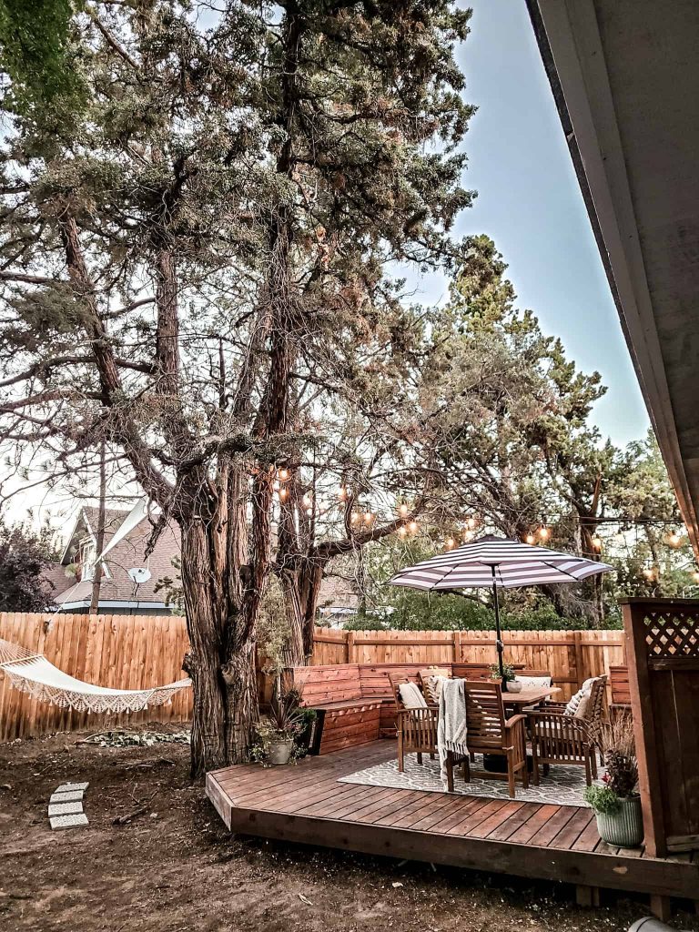 The view of our cozy patio space from our backyard; we have a wood deck with cozy dining table and chairs, bench seats, and string lights hanging from the trees. We also have a hammock swing/