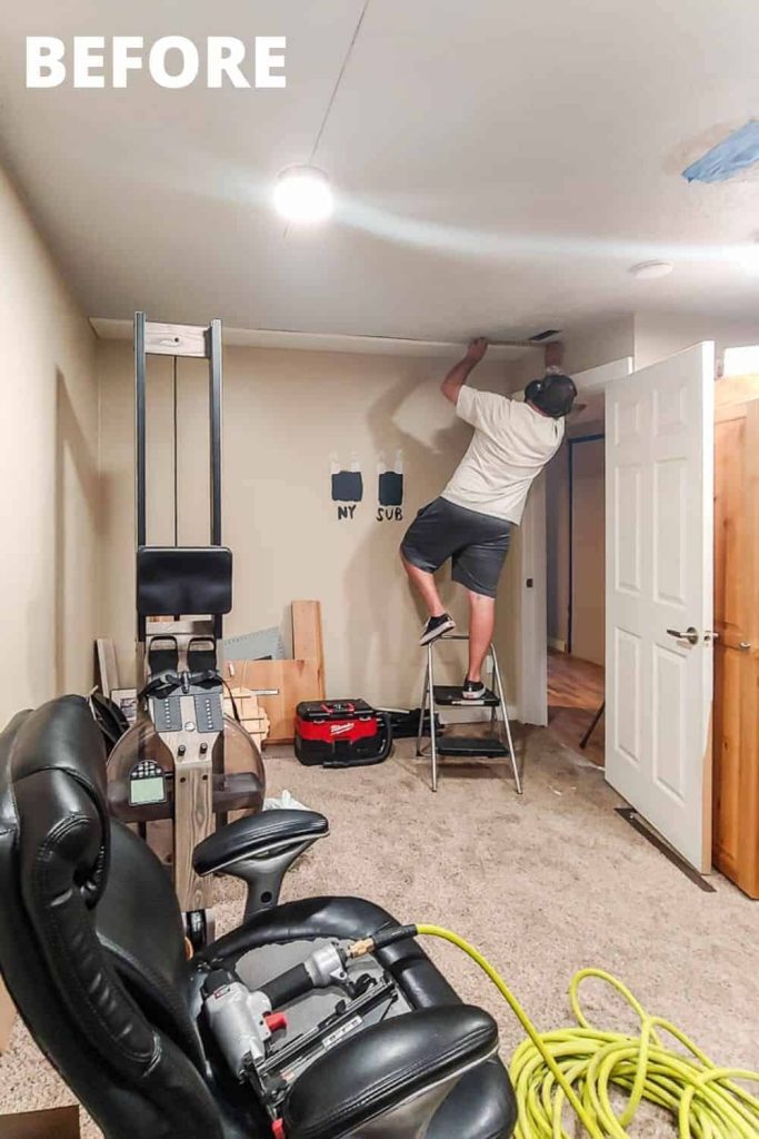 A picture of a cluttered room willed with random items like workout equipment, and office chair, and construction tools. A man stands on a step stool attaching wood to the ceiling.