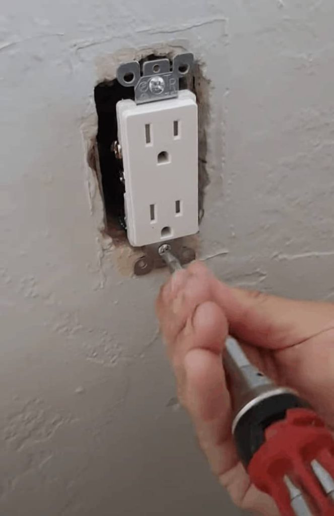 A person uses a screwdriver to attach a brand new white electrical outlet to a white wall.