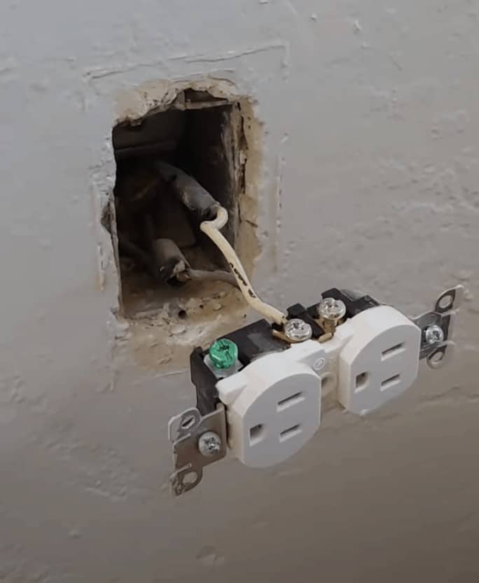 An old white electrical outlet sticks out from a hole in the wall, connected by two electrical wires.