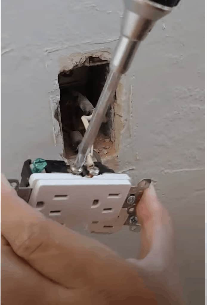 A person uses a screwdriver to unscrew the connection wires from an old electrical outlet hanging from a white wall.