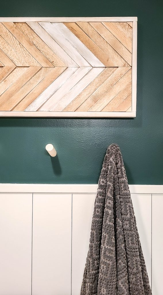 wooden wall art piece on a dark teal wall with shiplap walls and wood dowels underneath it with a towel