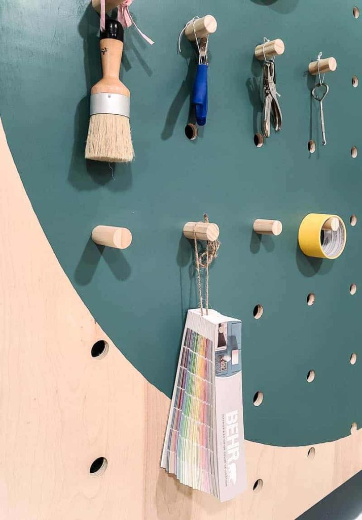 Shorter dowel rods on the giant pegboard wall hold DIY supplies like a paint colors sample book, paint brushes, crafting supplies, and tape.