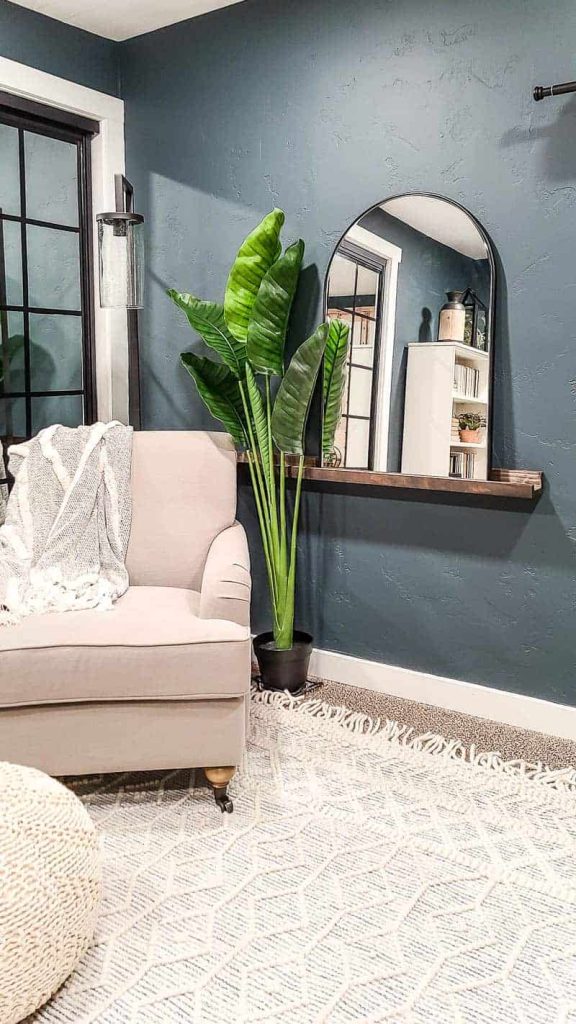 arched mirror with black frame on a dark wood shelf ledge next to a faux tropical plant, boho textured area rug with tassels, and white linen chair with dark teal walls