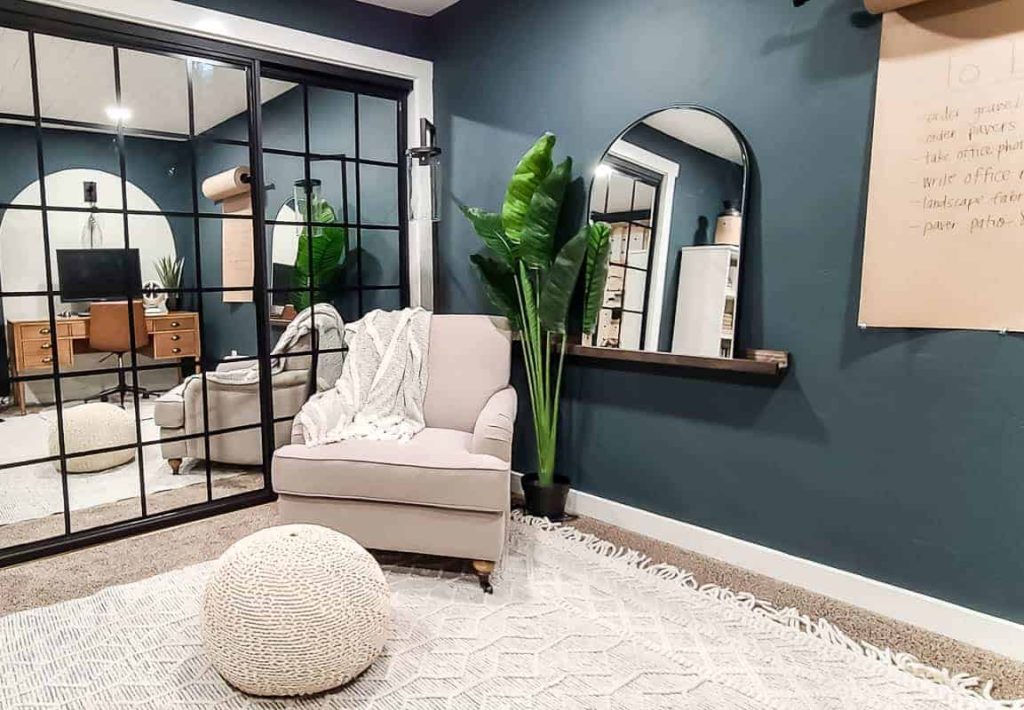 shows the corner of the dark teal office with a white arm chair, arch mirror on ledge shelf, plant with white boho rug, closet doors with mirror 