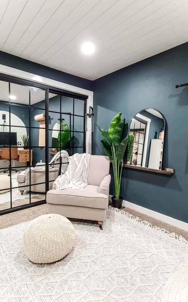 shows a mirror with rounded top and black frame on a dark wood shelf ledge next to a plant and white chair and macrame floor poof foot rest with dark teal walls with mirrored closet doors in background and white shiplap on ceiling 