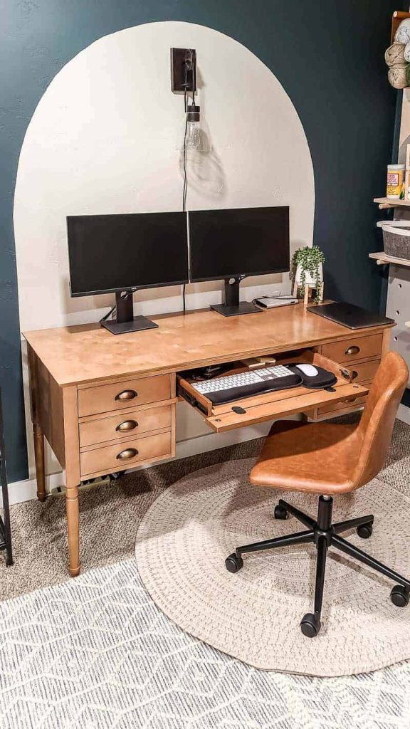 wood desk with two computer monitors and a leather task chair and white boho rug and dark teal walls 