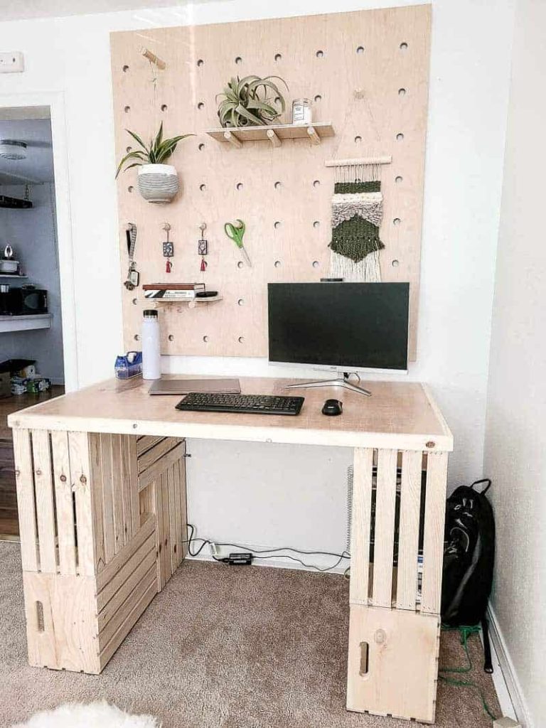 DIY wood crate desk with a computer monitor on it and a wood plywood pegboard wall above it