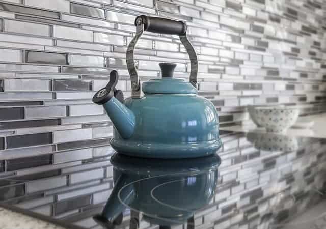 A teal blue tea kettle sits on a flat-top stove, with a gray and white glass tile backsplash.