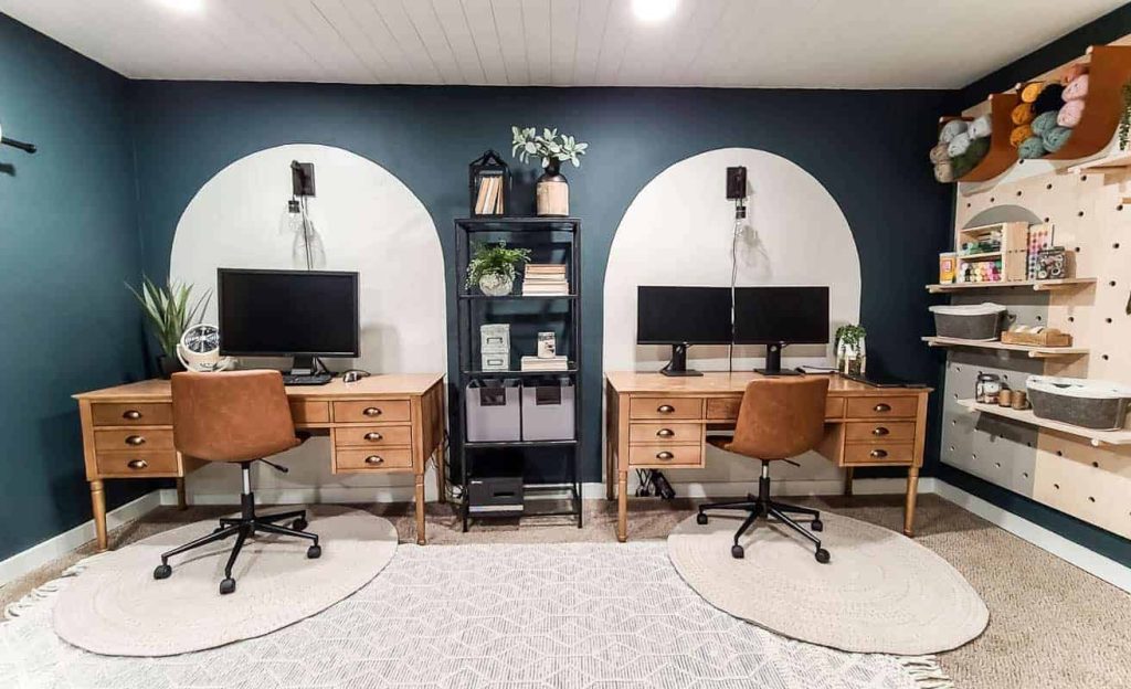 shows a dark green and white office space with two wooden desks, painted arches on wall, boho area rug and leather task chairs and a giant wood pegboard