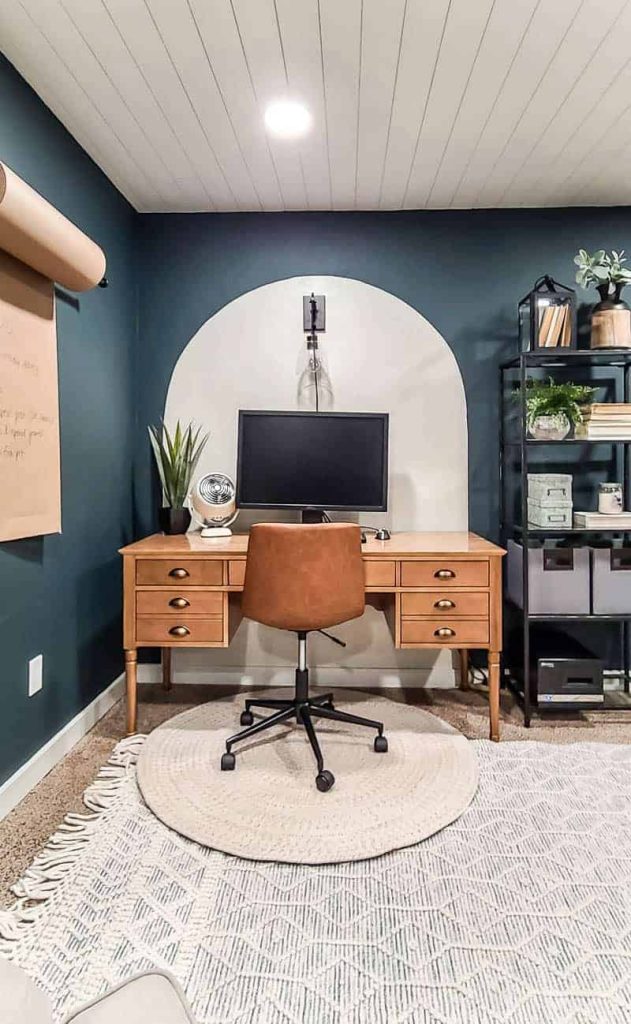 shows a white painted arch backdrop with a dark teal wall with a wood desk and leather chair with white rug with shiplap ceiling and industrial pully wall sconce light above desk and computer screen 