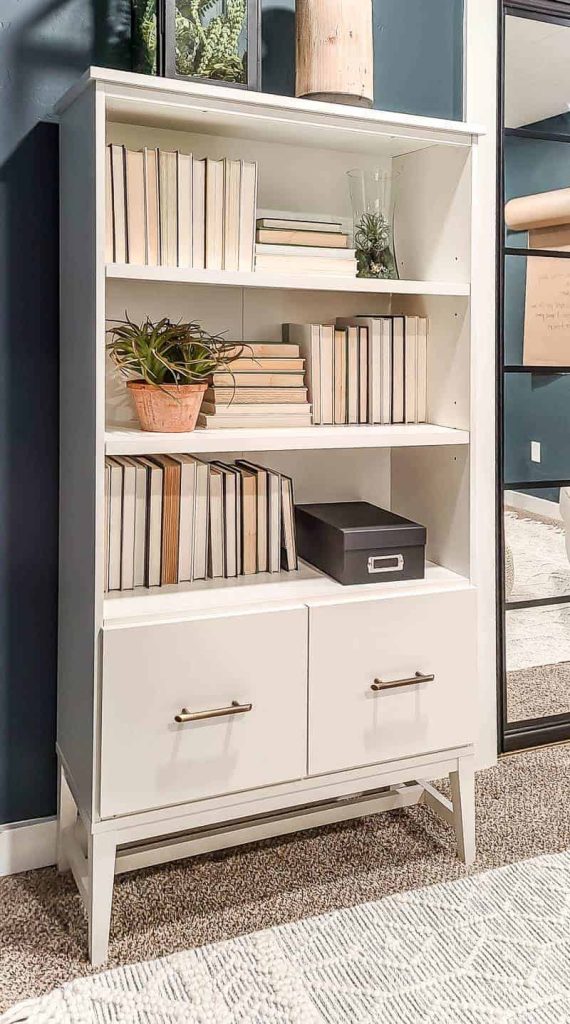 shows a white bookcase with 2 drawers for filing cabinet with books and plants with dark teal walls in home office