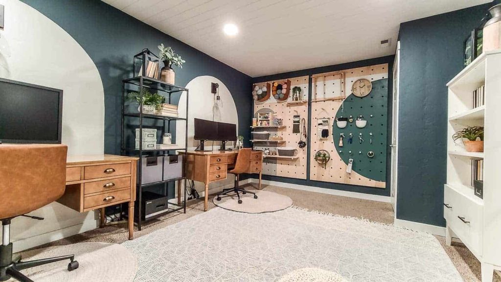 dark painted walls in home office with two wood desks and leather chairs with a giant wood pegboard wall with craft supplies and a white bookcase