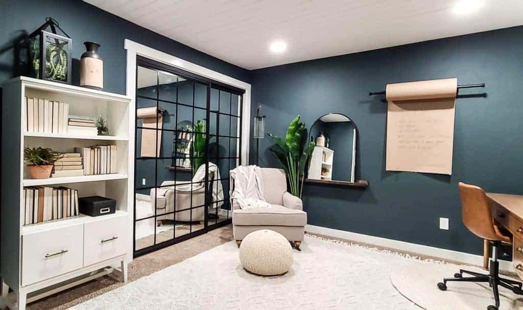 shows another corner of the dark green walls in home office with white shiplap ceiling and tan linen arm chair, arch mirror,  wall mounted paper roller and white bookcase 
