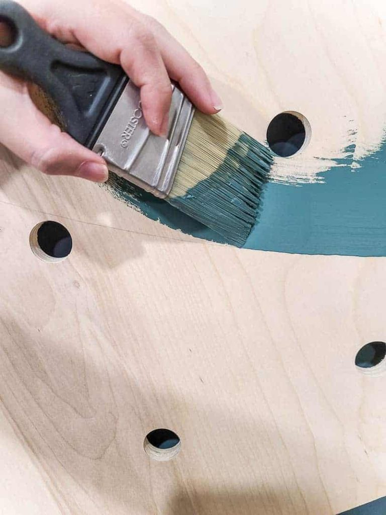 Using a paint brush and a teal blue paint to paint the bare wood of our giant pegboard wall.
