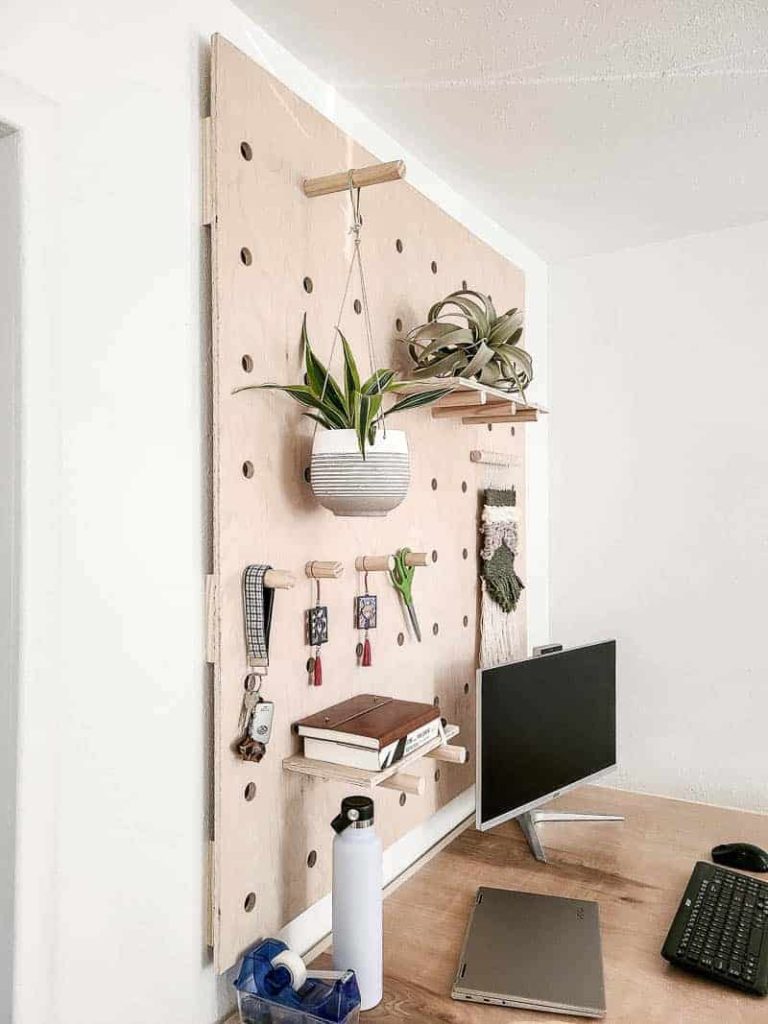 shows the top side of the wood crate desk with plants and other stuff hanging from a wood peg wall and has a computer on the desk 