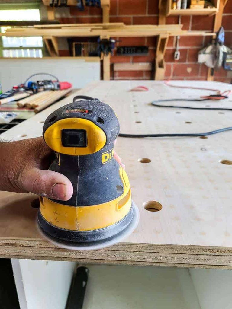 Using an orbital sander to sand down the surface of our pegboard wall, after drilling the pegboard holes.