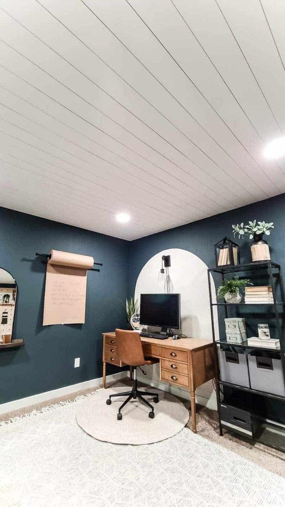 Modern boho office with dark blue wall sand wooden desk in corner with arch painted on the wall behind it with a large view of a white planked shiplap ceiling with recessed lights