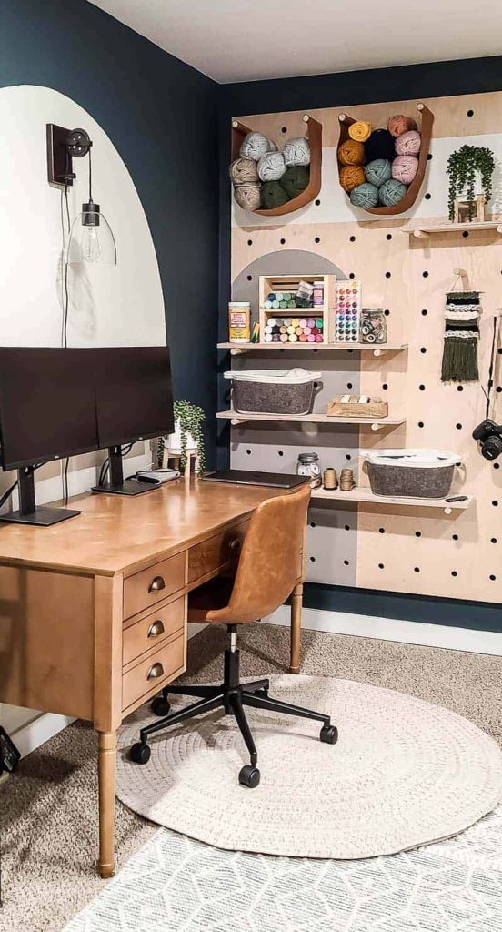 shows the giant wood pegboard with craft supplies next to a wood desk with a leather chair on a white rug with a white backdrop and dark teal walls