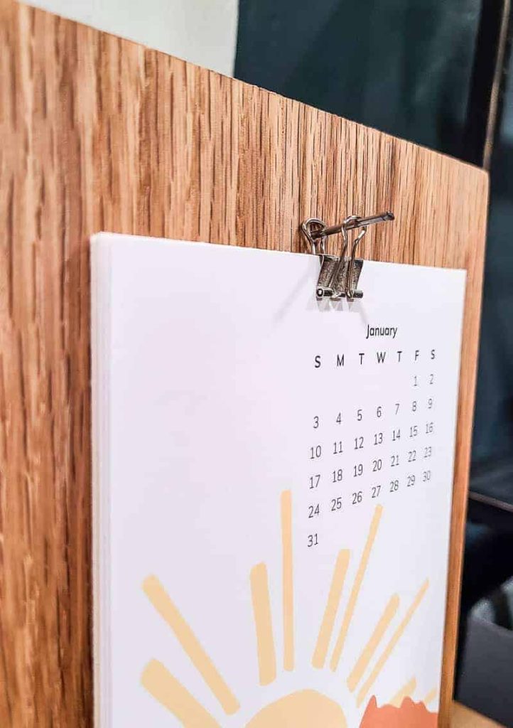 A closeup image of a January calendar pinned to a wooden desk calendar holder. The calendar features a boho style sun with golden beams.