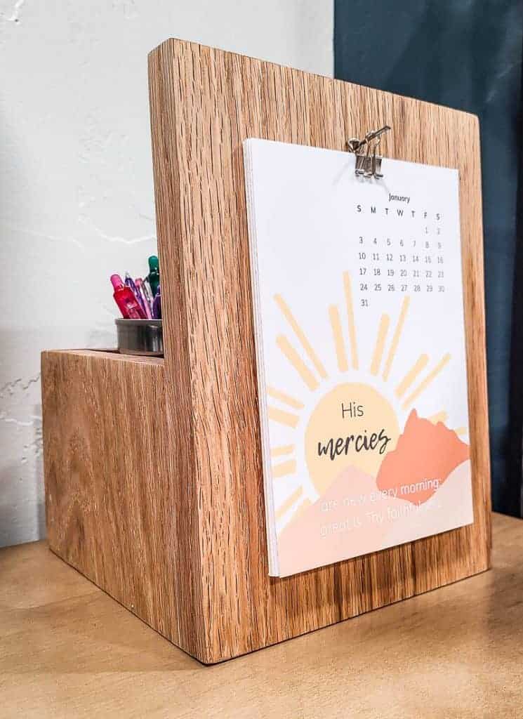 A close up front view of a DIY wooden desk calendar and pencil holder with a paper January calendar hanging on the front. The calendar features an illustration of a sun riding over mountains.