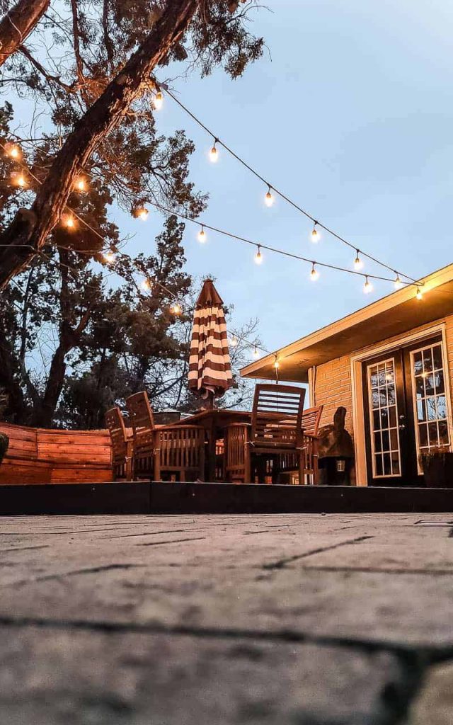 shows a view of the wood stained deck under edison bulb string lights in backyard with wooden table and striped umbrella