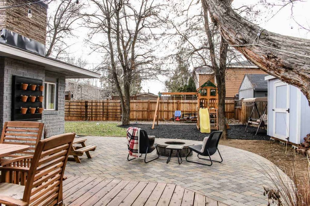 shows paver patio and fire pit with chairs and swing set in backyard with fence and grass shown in background
