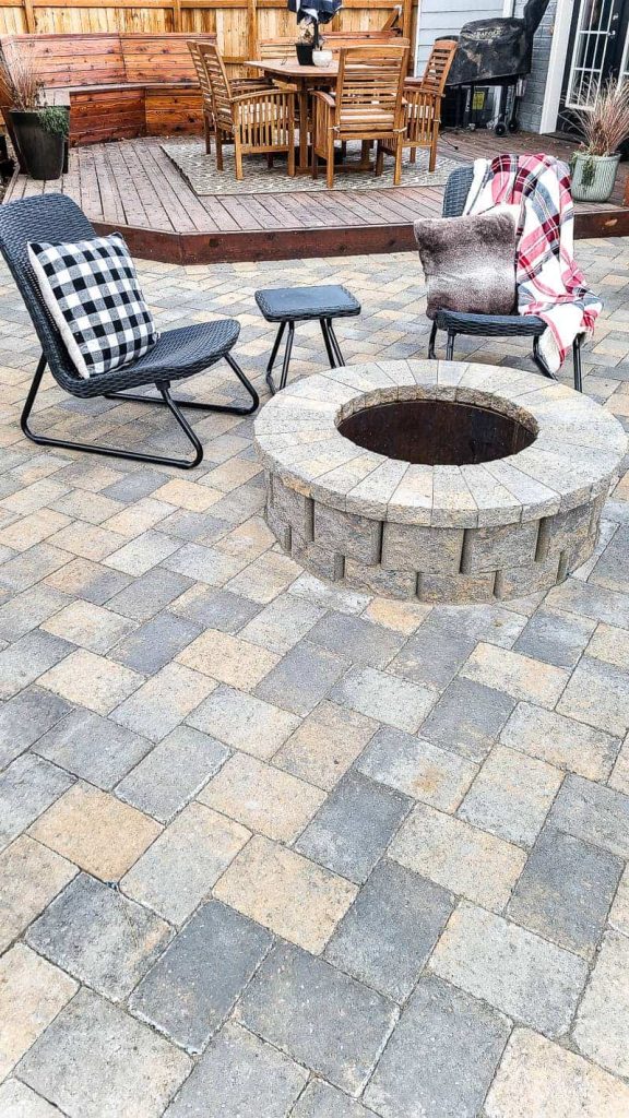 shows the finished round fire pit with metal ring and paver patio with chairs and deck with outdoor dining furniture in background