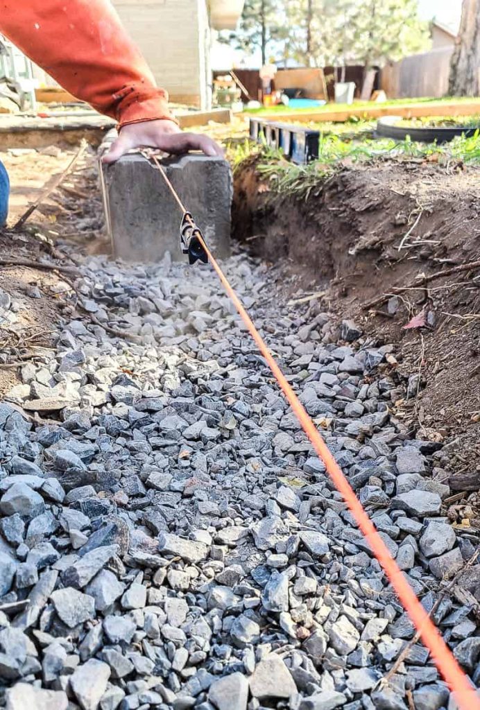 someone holds a retaining wall block steady in the gravel trench with a string line and level hanging over it