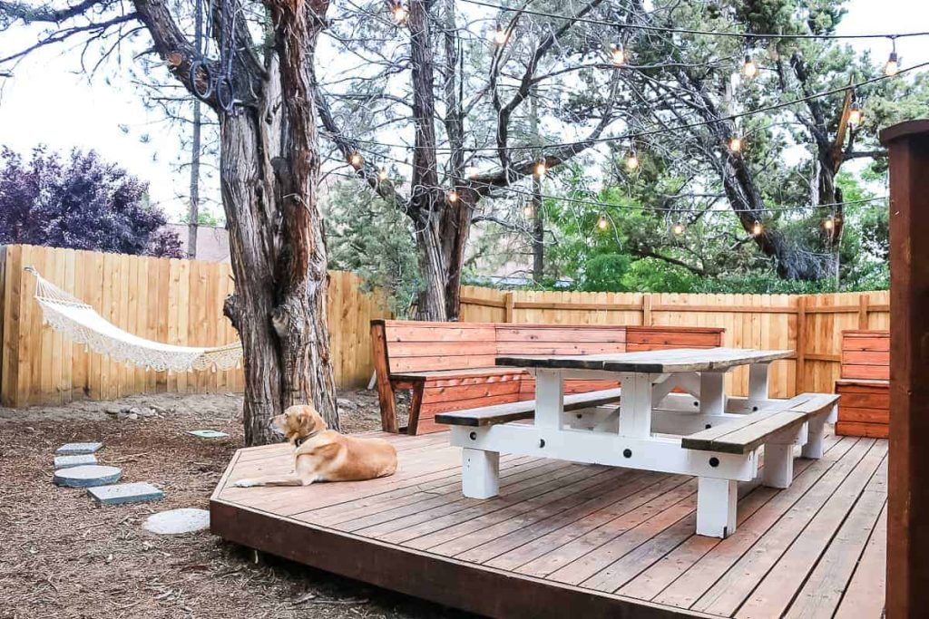 shows new backyard patio with large farmhouse wooden table, string lights overhead hanging from trees, fence in background and macrame hammock 