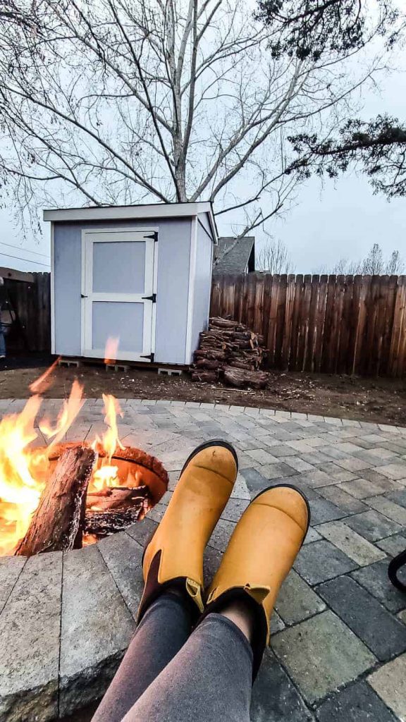 shows a shed in the background of the fireplace and yellow boots propped up on fire pit