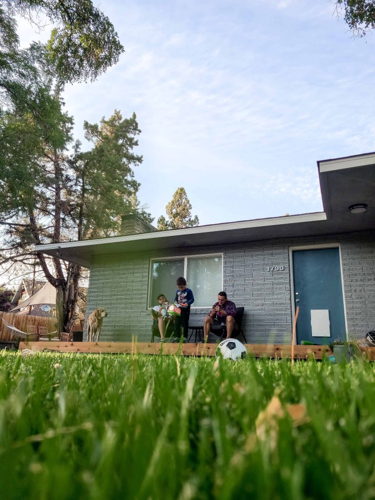 A low-to-the-ground image of a family sitting on a their back porch outside. A mom, dad, young boy and girl, and family dog sit in chairs. The sky is clear and blue, and the lush grass is healthy and green.