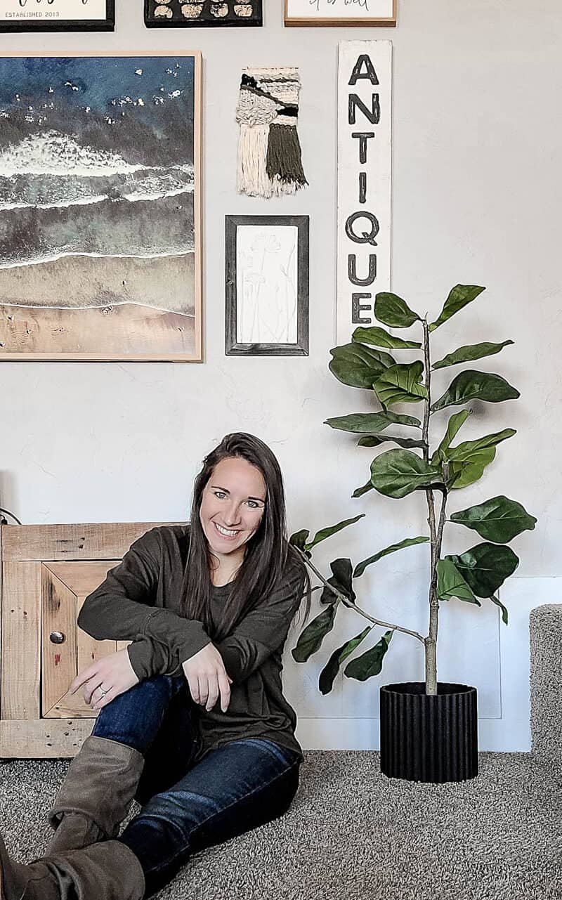 Girl sitting on ground next to faux fiddle leaf fig tree with black fluted planter pot cover on the base 