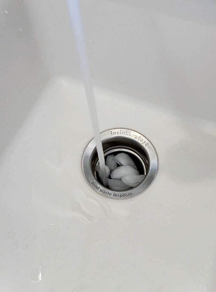 A steady stream of running water melts ice cubes and salt down a garbage disposal.