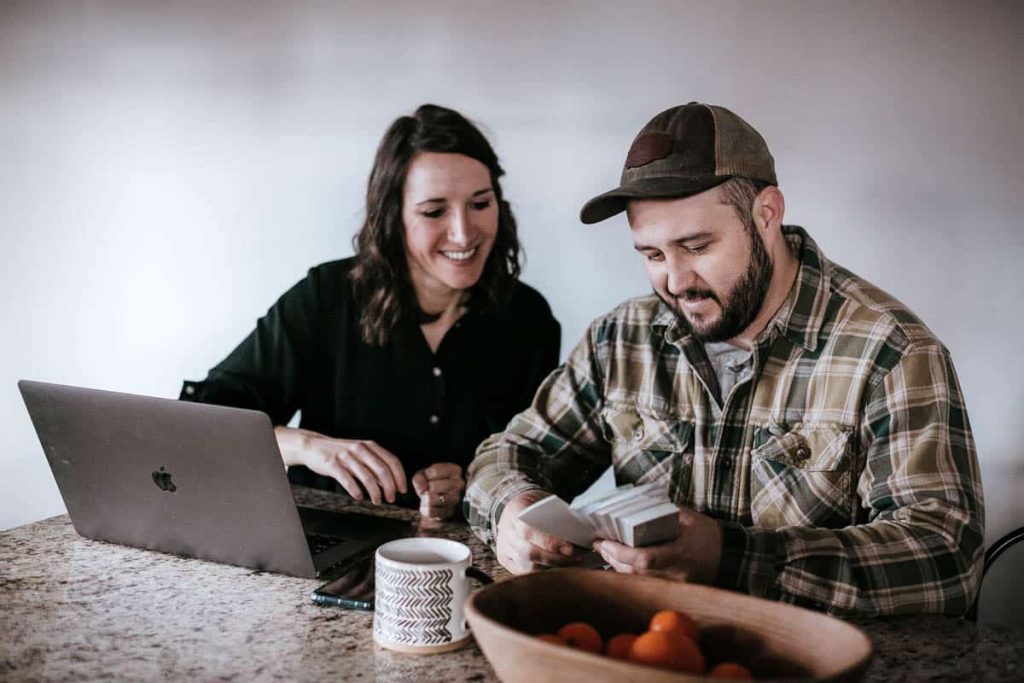 Husband and wife sitting at kitchen counter with laptop open and looking through paint samples while planning house DIY projects