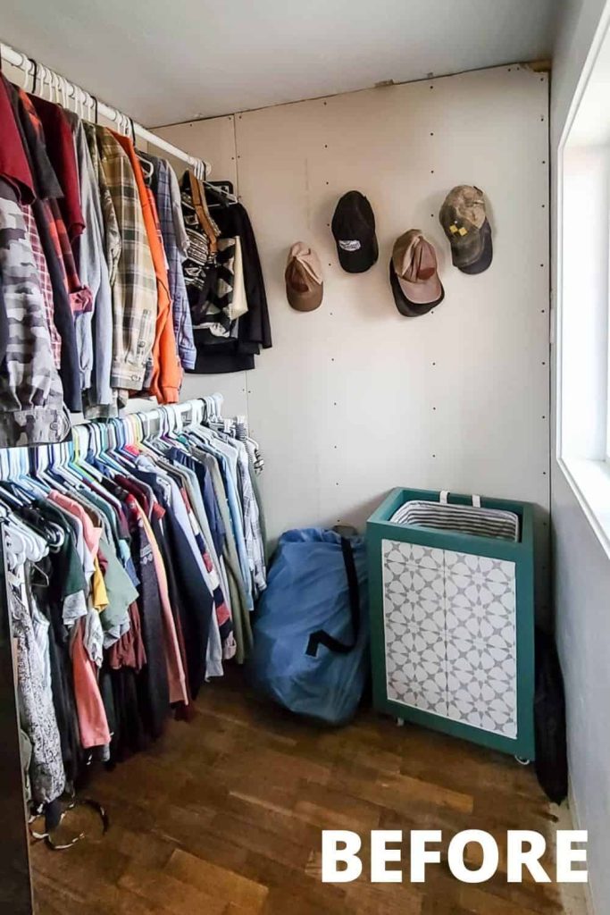 Our unfinished walk-in closet features plain drywall boards and clothing poles filled with hangers of clothing, and a laundry hamper in the corner.