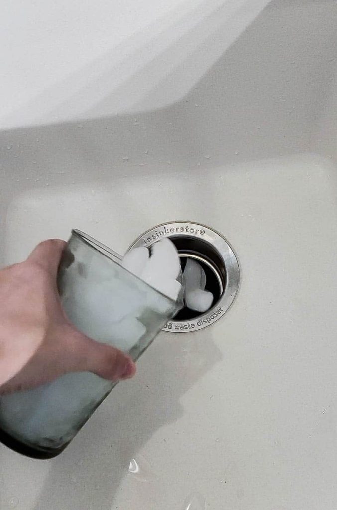 A woman pours a cup full of ice cubes Into a garbage disposal drain.