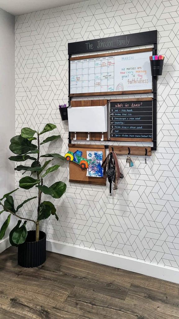 Our family command center, hanging on the wall in our mudroom. The command center features elements like a whiteboard calendar, mail holder, and key hooks for easy organization. The board conveniently hangs on a wallpapered wall next to a tall potted plant.