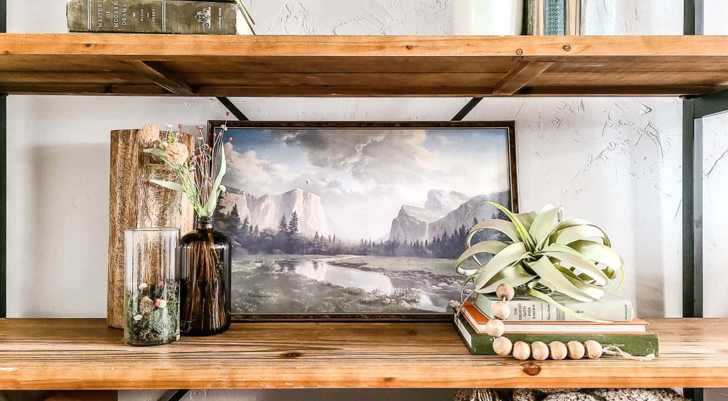 A wooden shelf decorated with vases filled with moss, rocks, and dried spring florals, a painting depicting a scenic mountain landscape, and a stack of vintage books.
