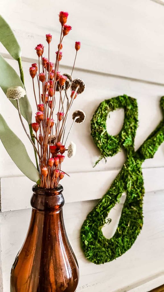 A close up side angle view of letters cut out from sheet moss, spelling out the word "grace" attached to a wooden serving tray, next to a vase filled with dried florals.
