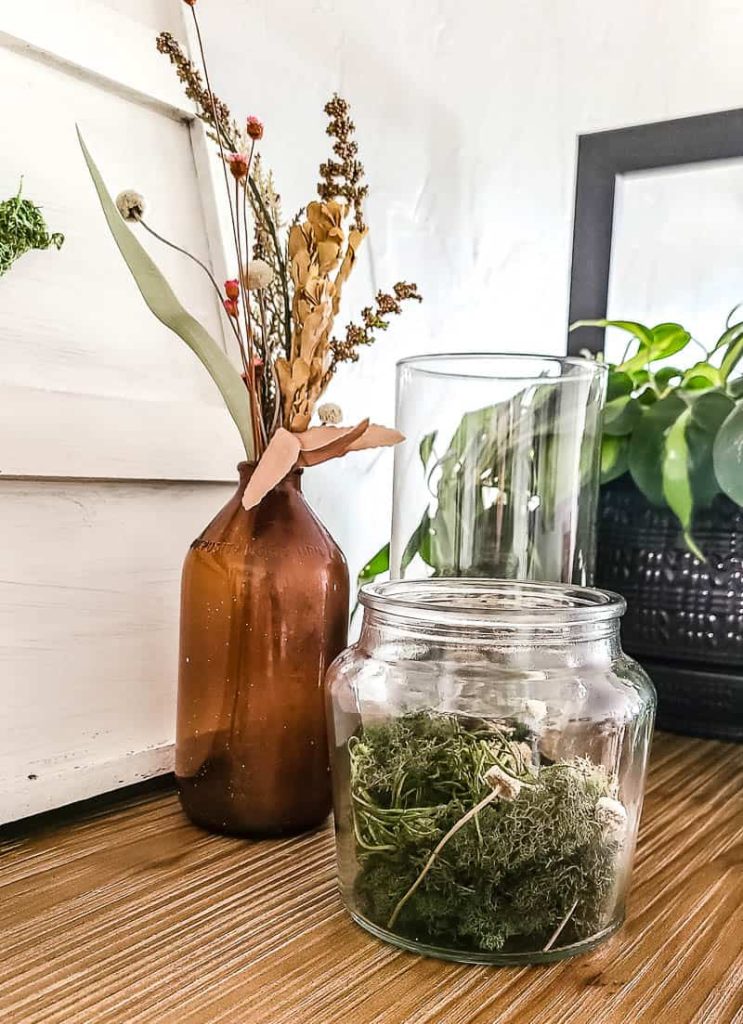 A group of glass and amber vases filled with faux moss, stones, and dried flowers on a wooden bookshelf.