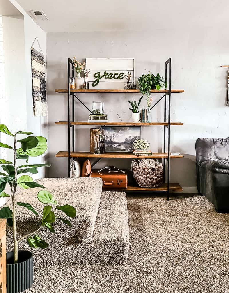 An industrial style wood and metal shelf against a white wall in a living room. The shelved are lined with plants, flowers, pictures, vintage books, and more boho style decor pieces.